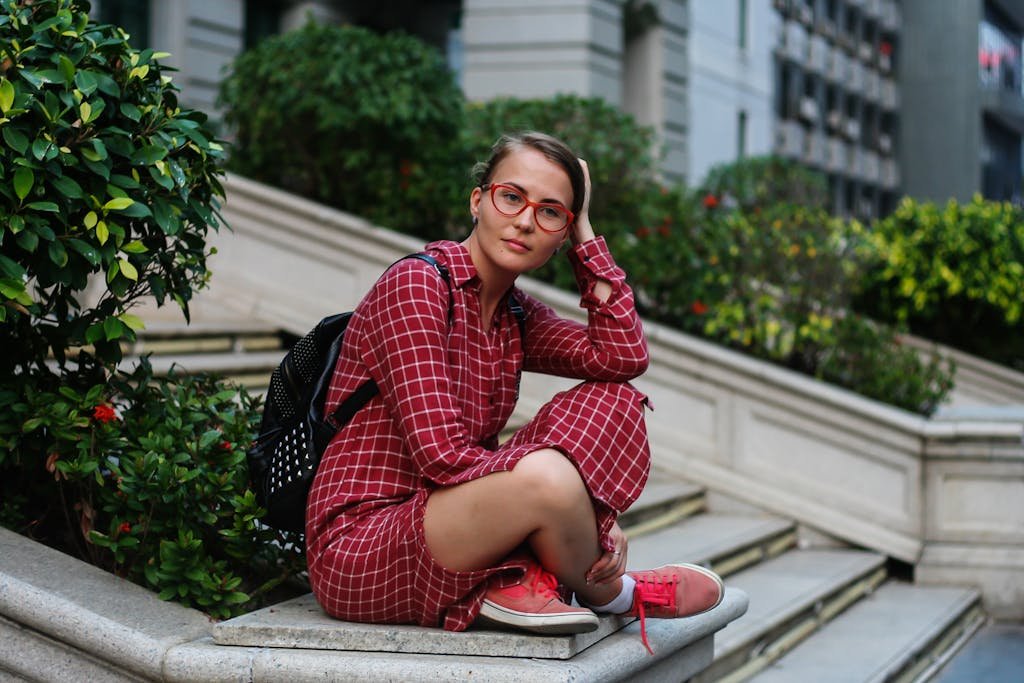 A young woman wearing a red plaid dress and glasses sits on outdoor stairs surrounded by greenery.