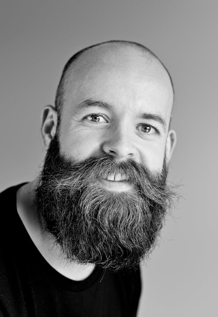 Black and white portrait of a smiling bald man with a large beard in a studio setting.