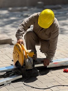 A construction worker wearing a yellow helmet cuts metal using a circular saw outdoors.
