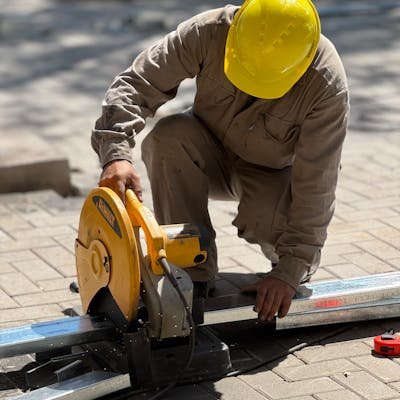 A construction worker wearing a yellow helmet cuts metal using a circular saw outdoors.
