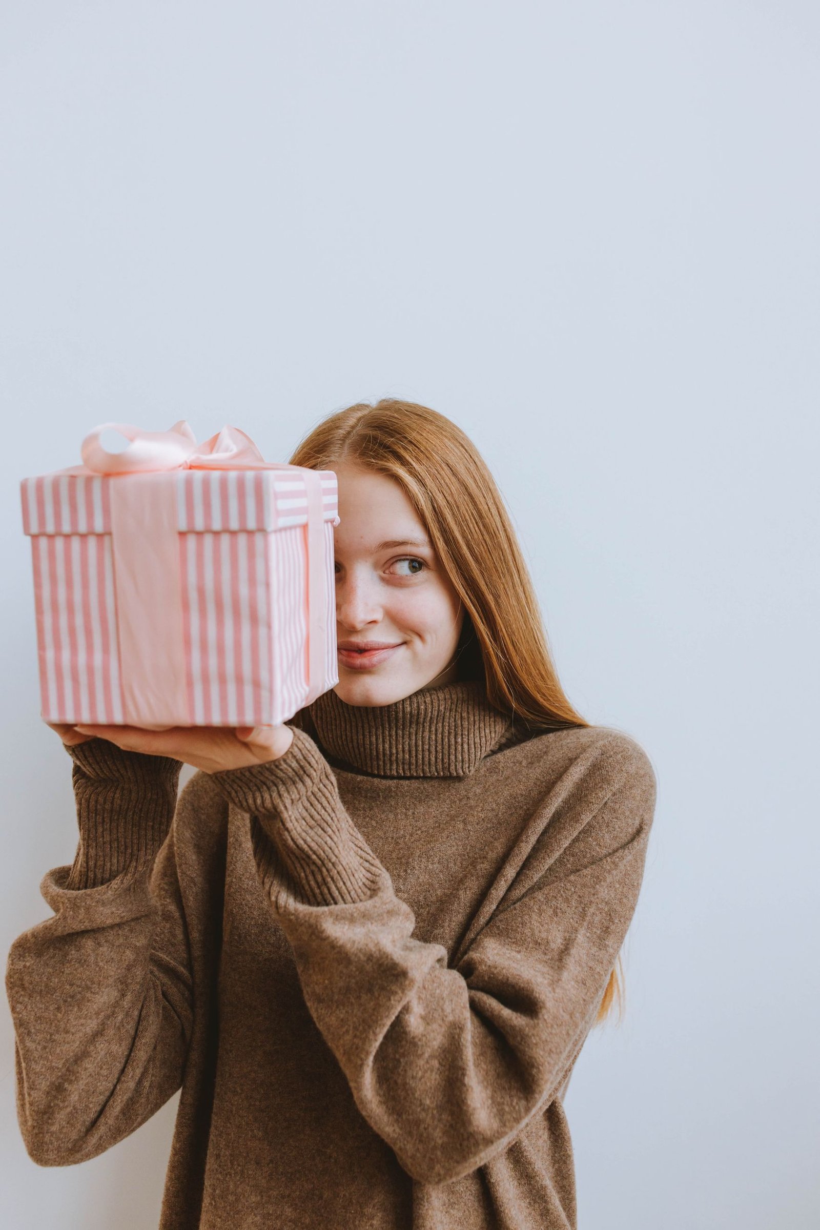 Caucasian woman in a cozy sweater holds a pink gift box, smiling in a studio setting.